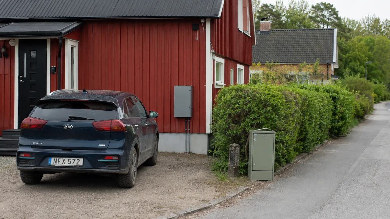 Blå elbil vid ett rött trähus med elskåp på fasaden och vid gatan.