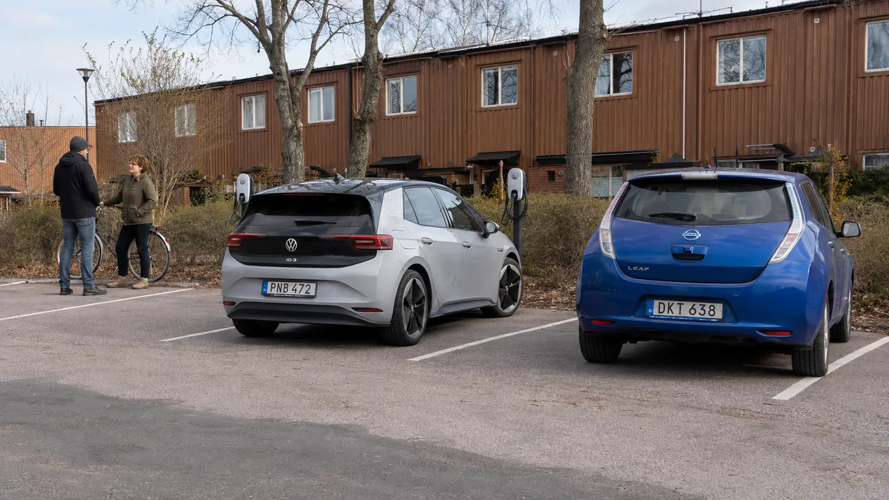 Elbilar och laddstolpar vid en gemensam radhusparkering där två boende samtalar.